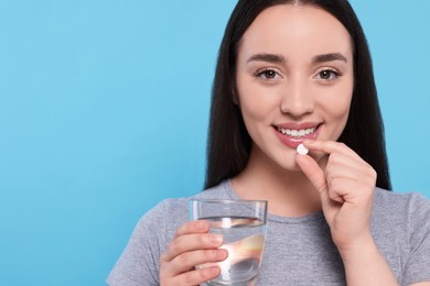 Beautiful young woman with glass of water taking pill on light blue background, space for text