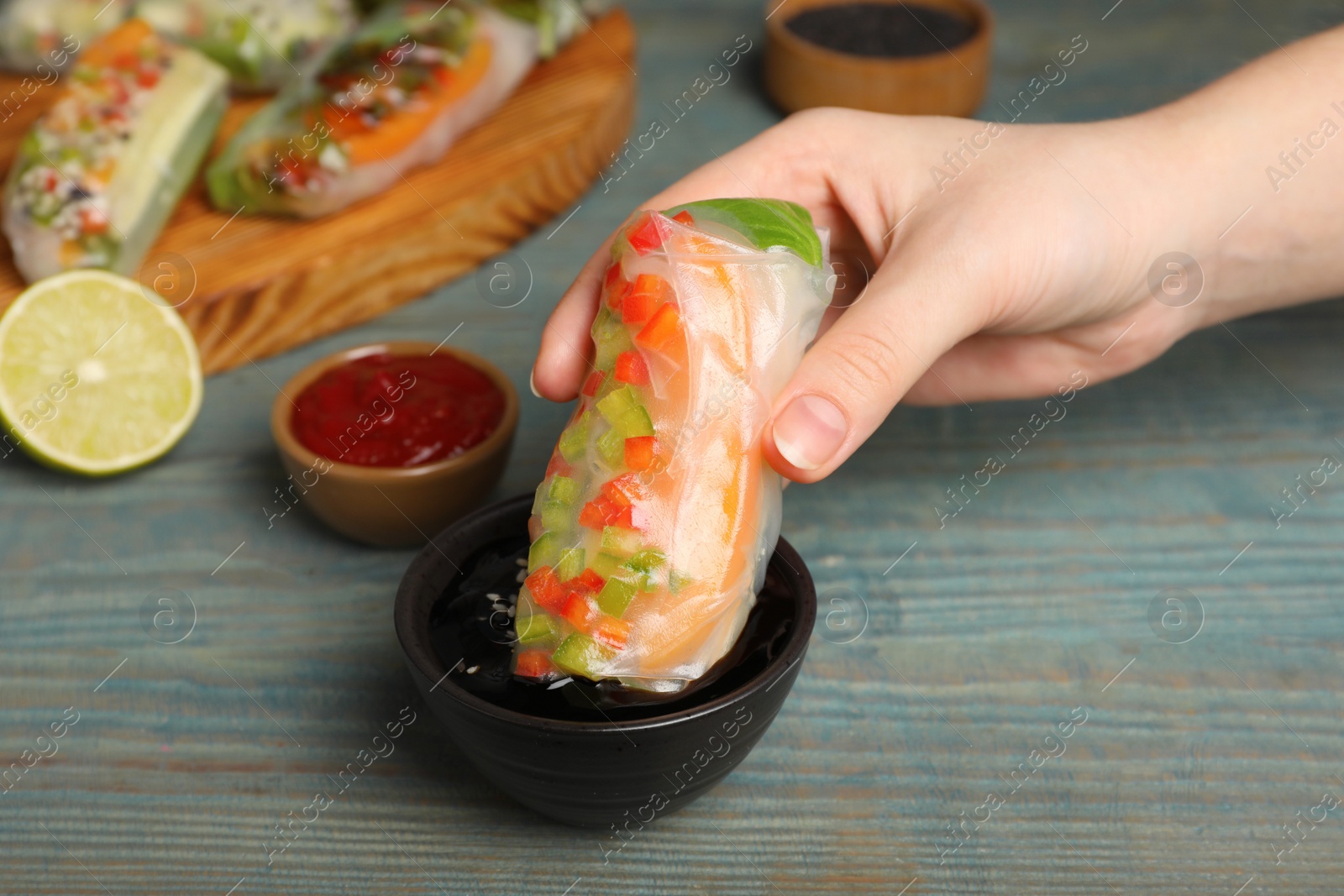 Photo of Woman dipping delicious roll wrapped in rice paper into soy sauce at blue wooden table, closeup