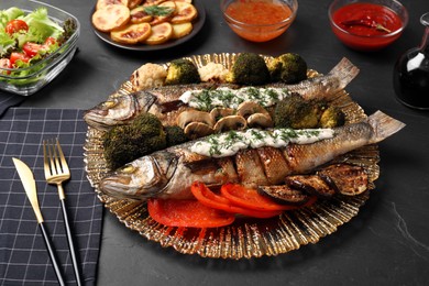 Photo of Plate with delicious baked sea bass fish and sauce served on dark table