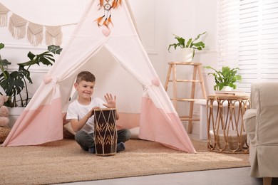 Photo of Cute little boy playing drum near toy wigwam at home