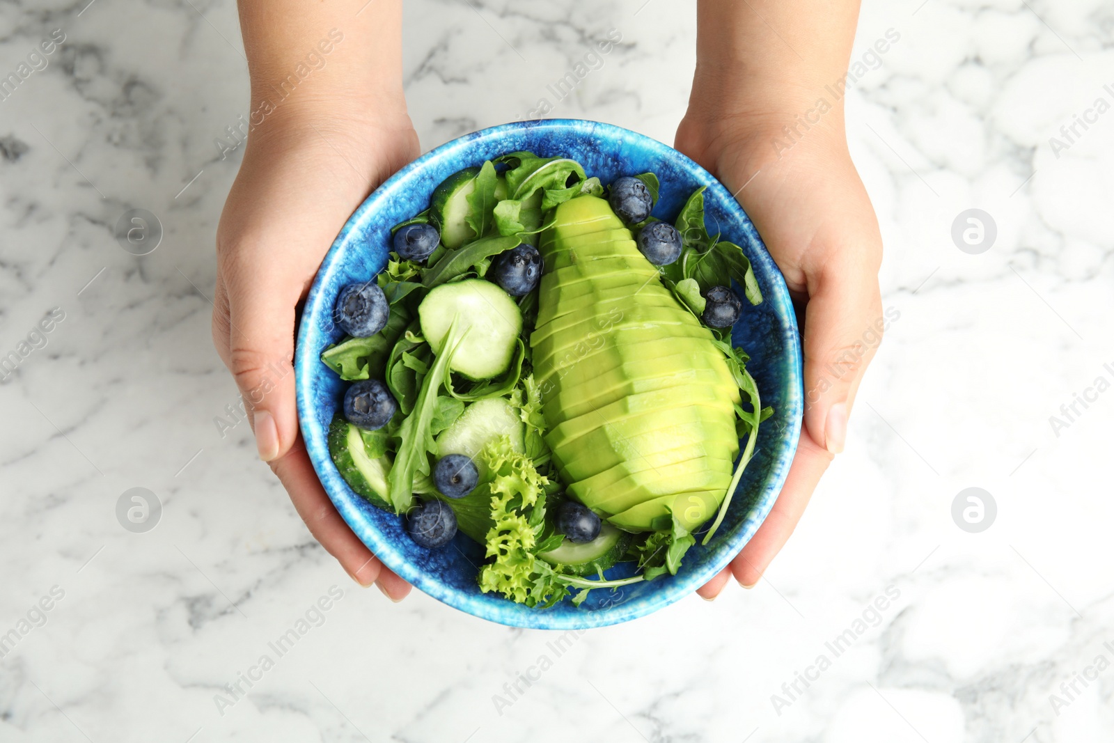 Photo of Woman holding bowl of avocado salad with blueberries over marble table, top view