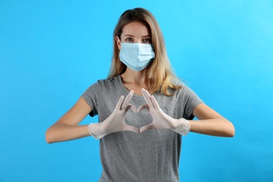 Young woman in medical gloves and protective mask making heart with hands on light blue background