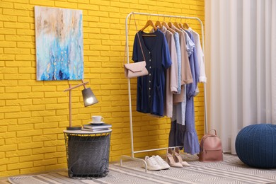 Rack with different stylish clothes and shoes near yellow brick wall in room