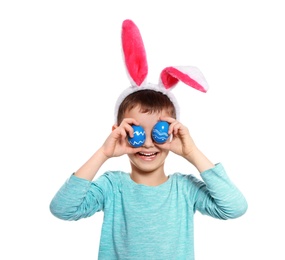 Little boy in bunny ears headband holding Easter eggs near eyes on white background