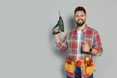 Young worker with power drill and tool belt on grey background. Space for text