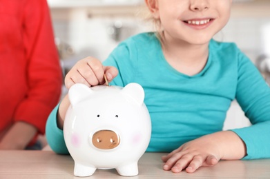 Cute girl putting coin into piggy bank at table, closeup. Saving money
