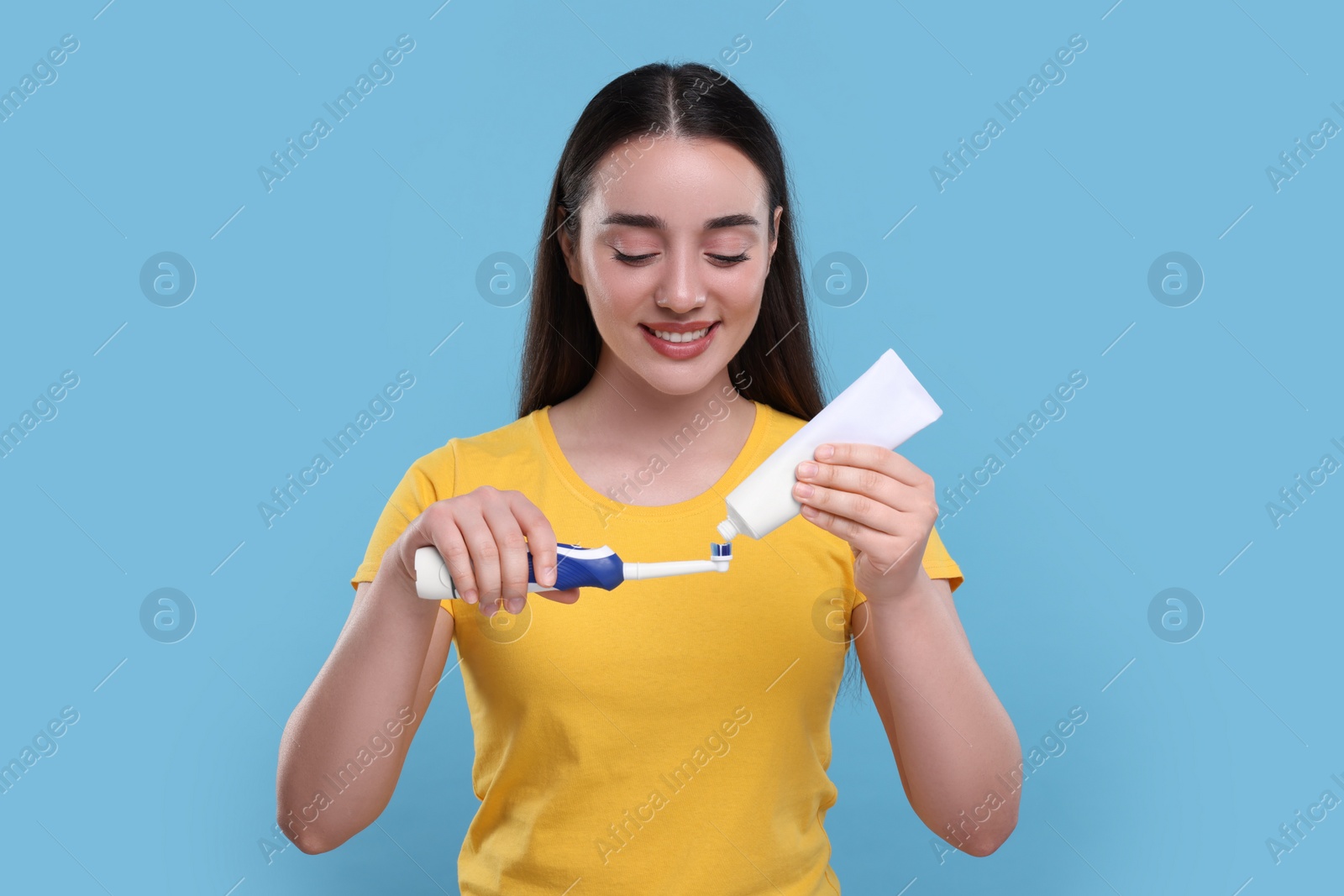 Photo of Happy young woman squeezing toothpaste from tube onto electric toothbrush on light blue background