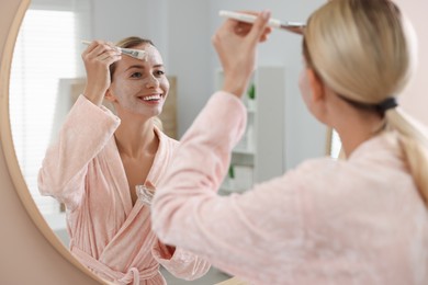 Woman applying face mask near mirror in bathroom. Spa treatments