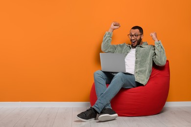 Smiling young man with laptop sitting on beanbag chair near orange wall, space for text