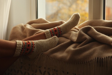 Photo of Woman in knitted socks relaxing on plaid near window at home, closeup