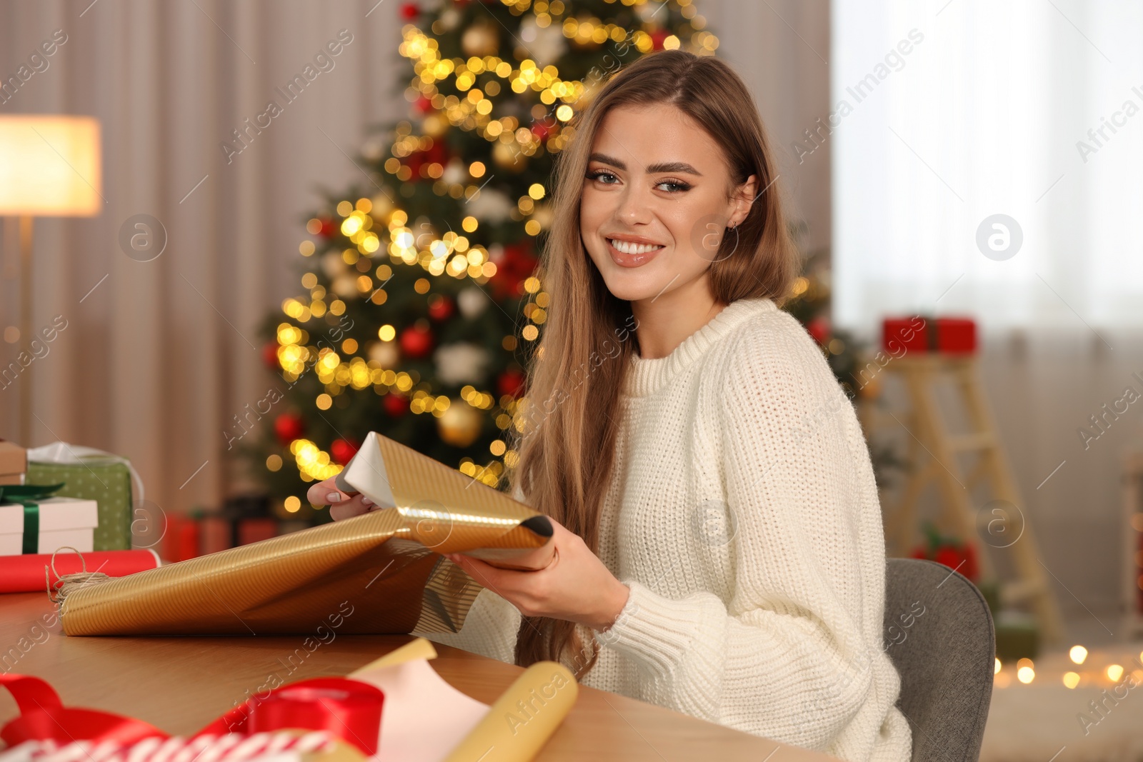 Photo of Beautiful young woman with wrapping paper at table in room. Decorating Christmas gift