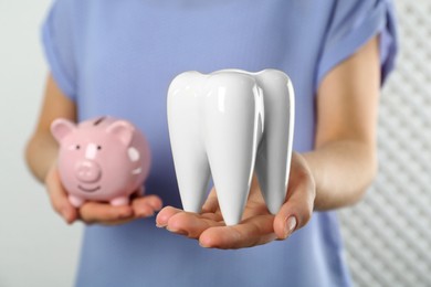 Woman holding ceramic model of tooth and piggy bank on light background, closeup. Expensive treatment
