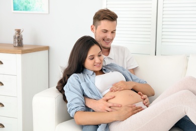 Photo of Young husband and his pregnant wife resting on sofa at home