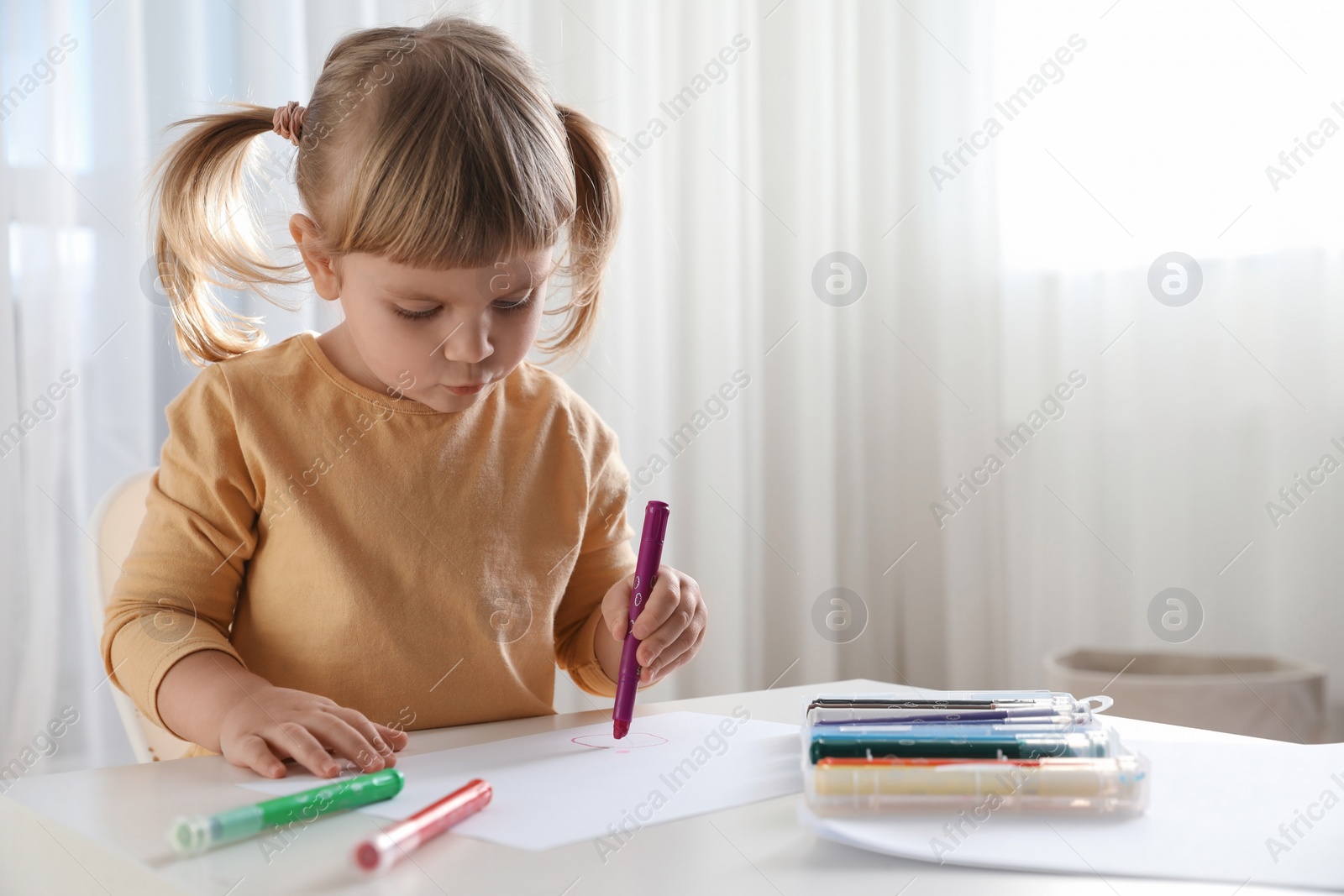 Photo of Cute little girl drawing with marker at white table indoors. Child`s art