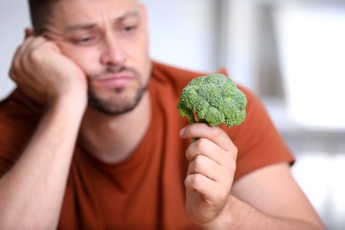 Unhappy man with broccoli on light background, closeup