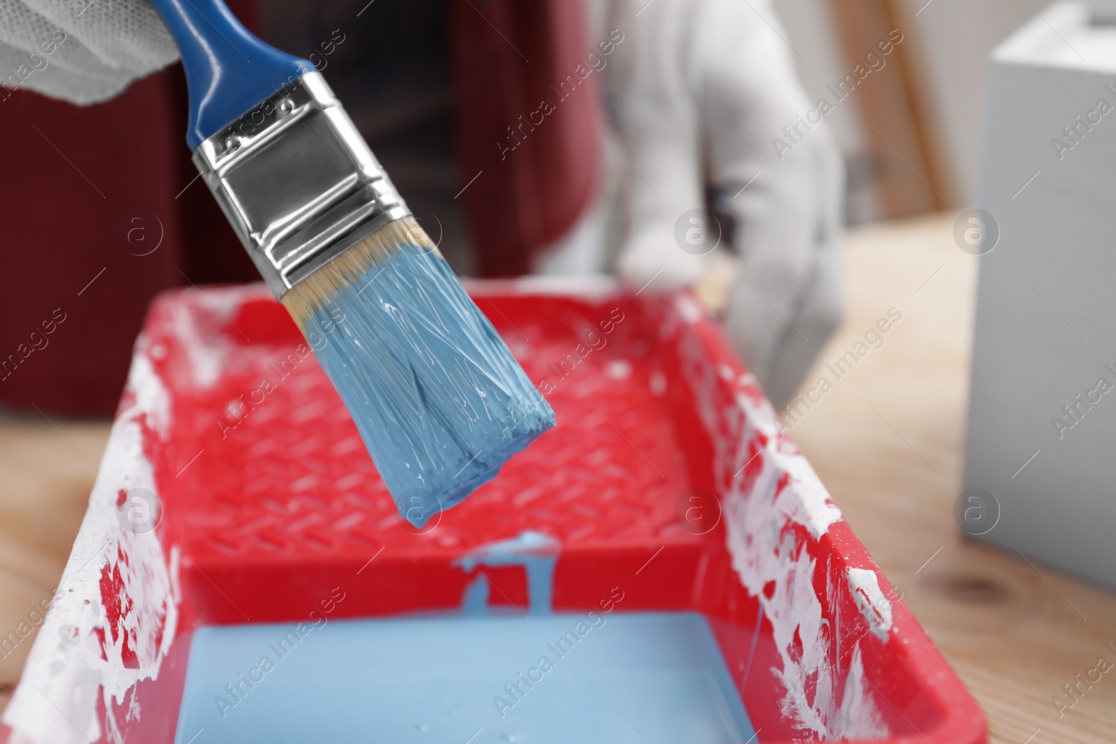 Photo of Woman taking light blue paint with brush from tray at wooden table, closeup