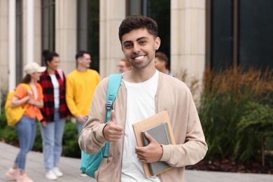 Photo of Students spending time together outdoors. Happy young man with notebooks showing thumbs up, selective focus
