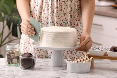 Photo of Woman using scraper to decorate cake at white marble table in kitchen, closeup