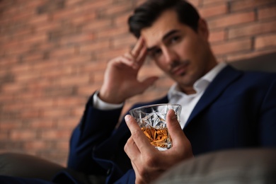 Young man with glass of whiskey near brick wall indoors