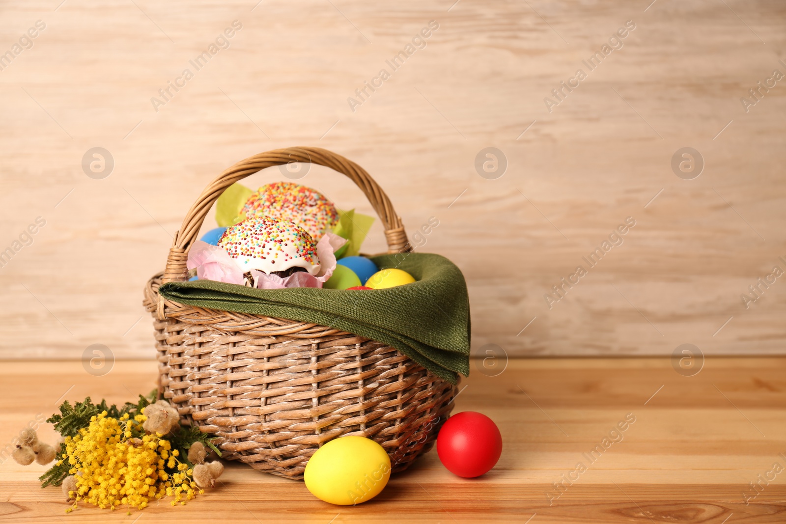 Photo of Basket with delicious Easter cakes, dyed eggs and flowers on wooden table. Space for text