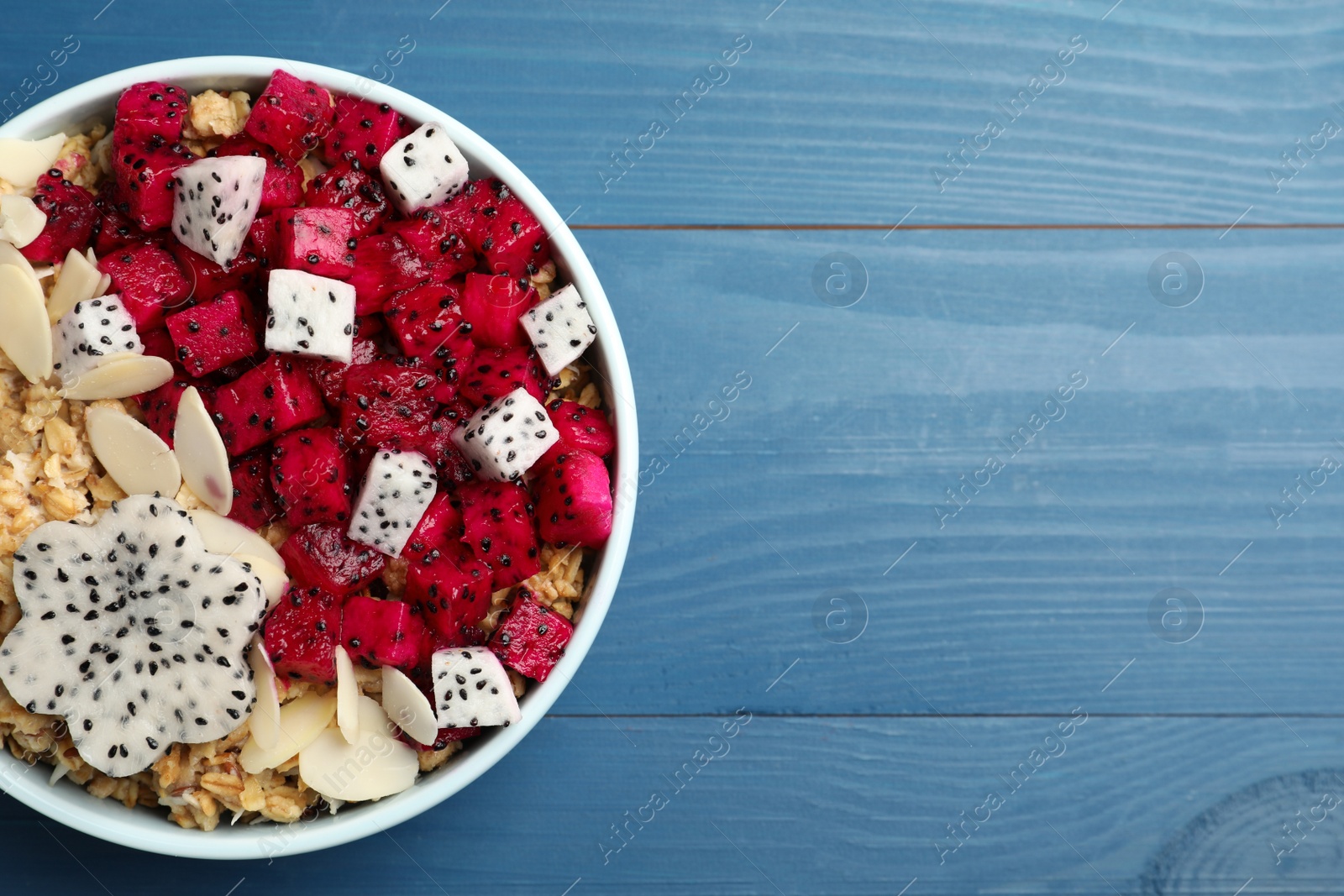 Photo of Bowl of granola with pitahaya and almond petals on blue wooden table, top view. Space for text