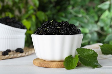 Fresh ripe black mulberries in bowl on white wooden table against blurred background