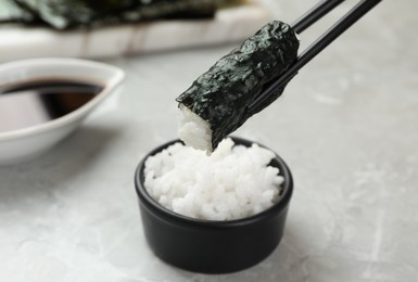 Photo of Chopsticks with cooked rice wrapped in nori sheet over light grey table, closeup