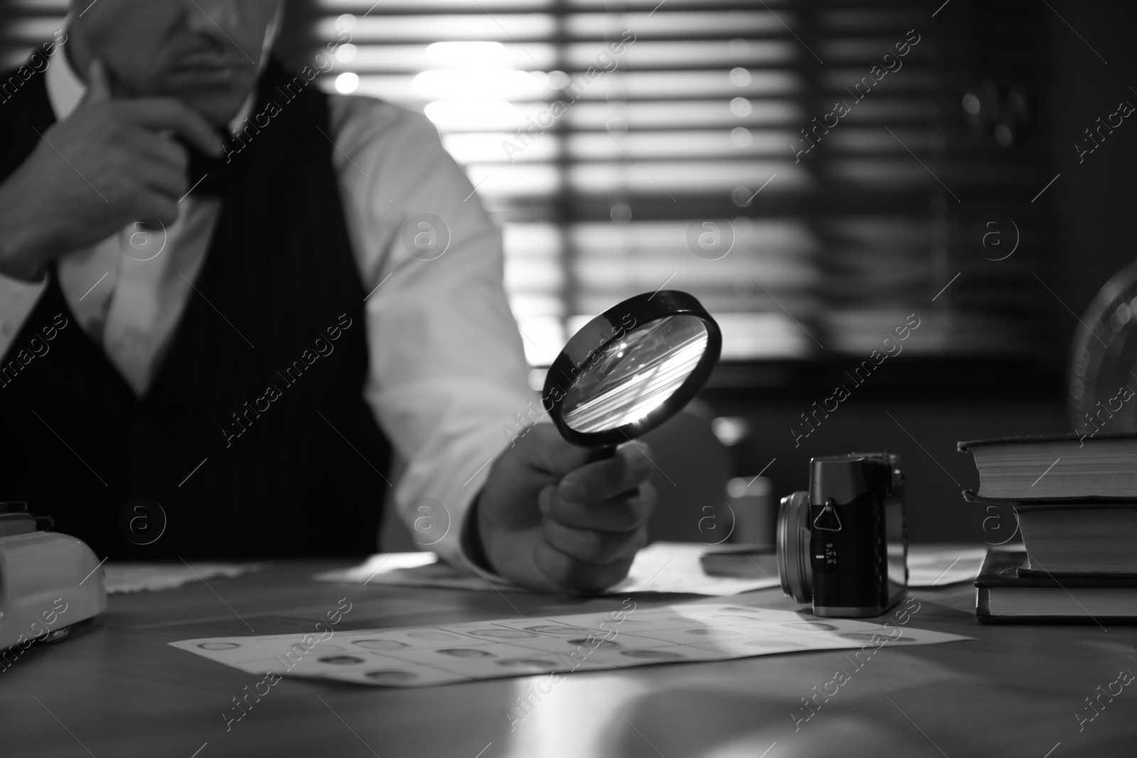 Photo of Old fashioned detective with magnifying glass working at table in office, closeup. Black and white effect