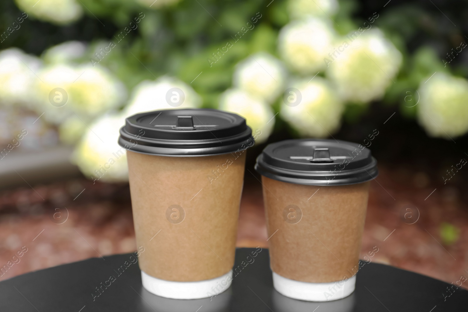 Photo of Cardboard cups with plastic lids on black table near flowers. Coffee to go