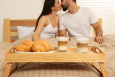 Photo of Happy couple kissing on bed at home, focus on wooden tray with breakfast