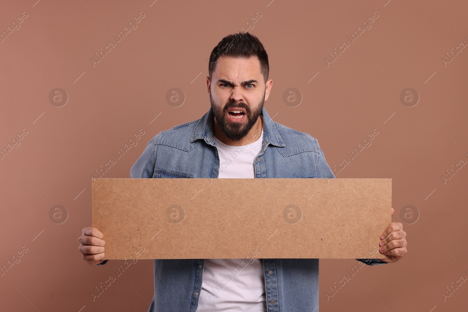 Photo of Angry young man holding blank cardboard banner on brown background, space for text