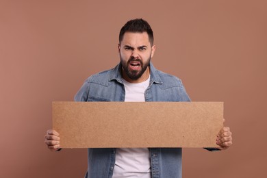 Photo of Angry young man holding blank cardboard banner on brown background, space for text