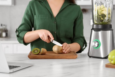 Young woman cutting kiwi for smoothie at white table in kitchen, closeup