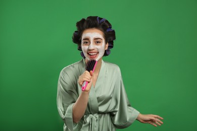 Photo of Happy young woman in silk bathrobe with hair curlers and facial mask singing into hairbrush on green background