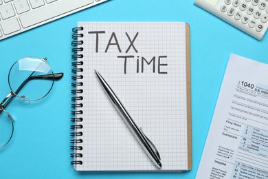 Photo of Notebook with words Tax Time, documents, computer keyboard, calculator and glasses on light blue background, flat lay