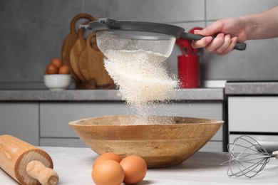 Woman sieving flour into bowl at white wooden table in kitchen, closeup