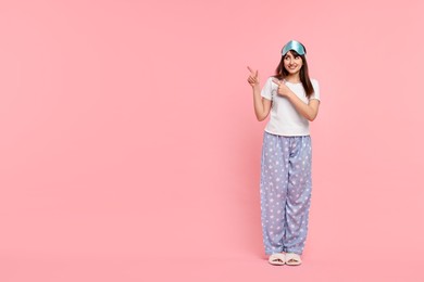 Happy woman in pyjama and sleep mask pointing at something on pink background, space for text