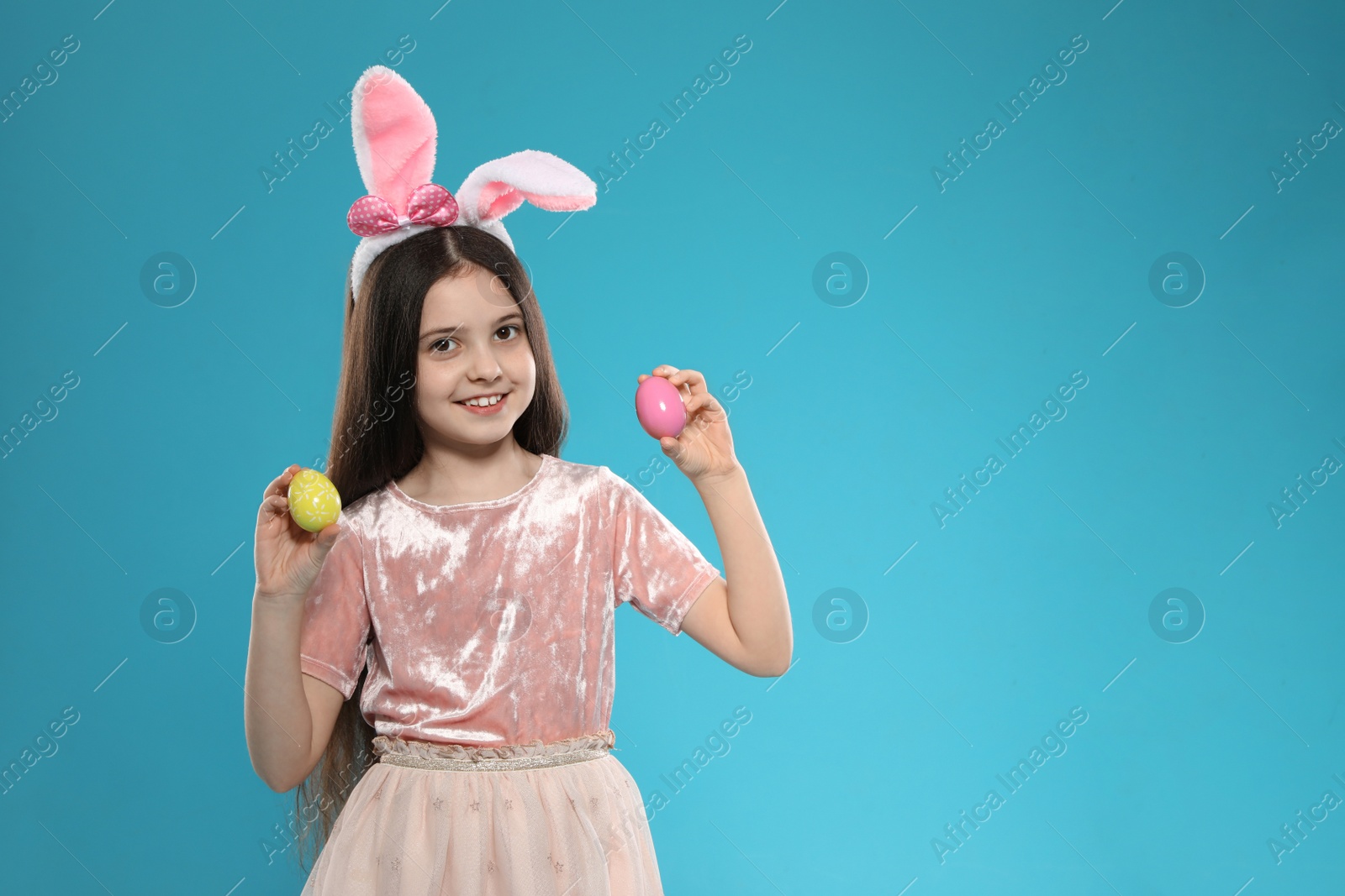 Photo of Little girl in bunny ears headband holding Easter eggs on color background, space for text