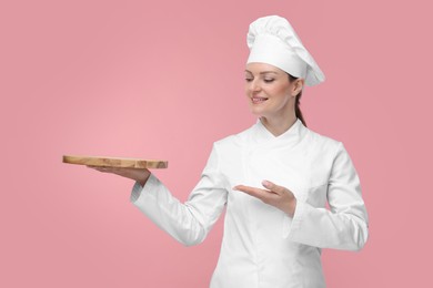 Photo of Happy chef in uniform holding empty wooden board on pink background