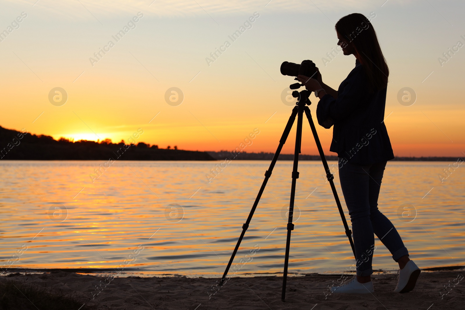 Photo of Young female photographer adjusting professional camera on tripod at sunset