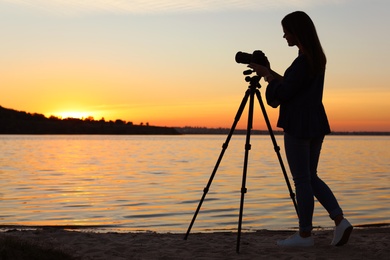 Young female photographer adjusting professional camera on tripod at sunset