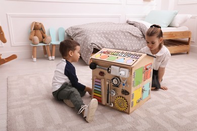 Photo of Little boy and girl playing with busy board house on floor in room