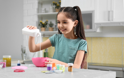 Photo of Cute little girl pouring glue into bowl at table in kitchen. DIY slime toy