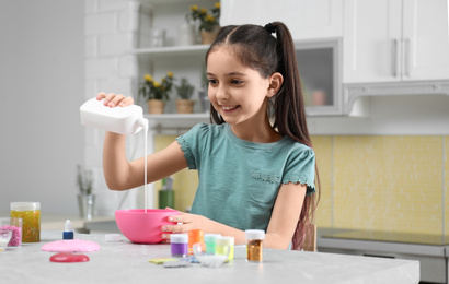 Cute little girl pouring glue into bowl at table in kitchen. DIY slime toy