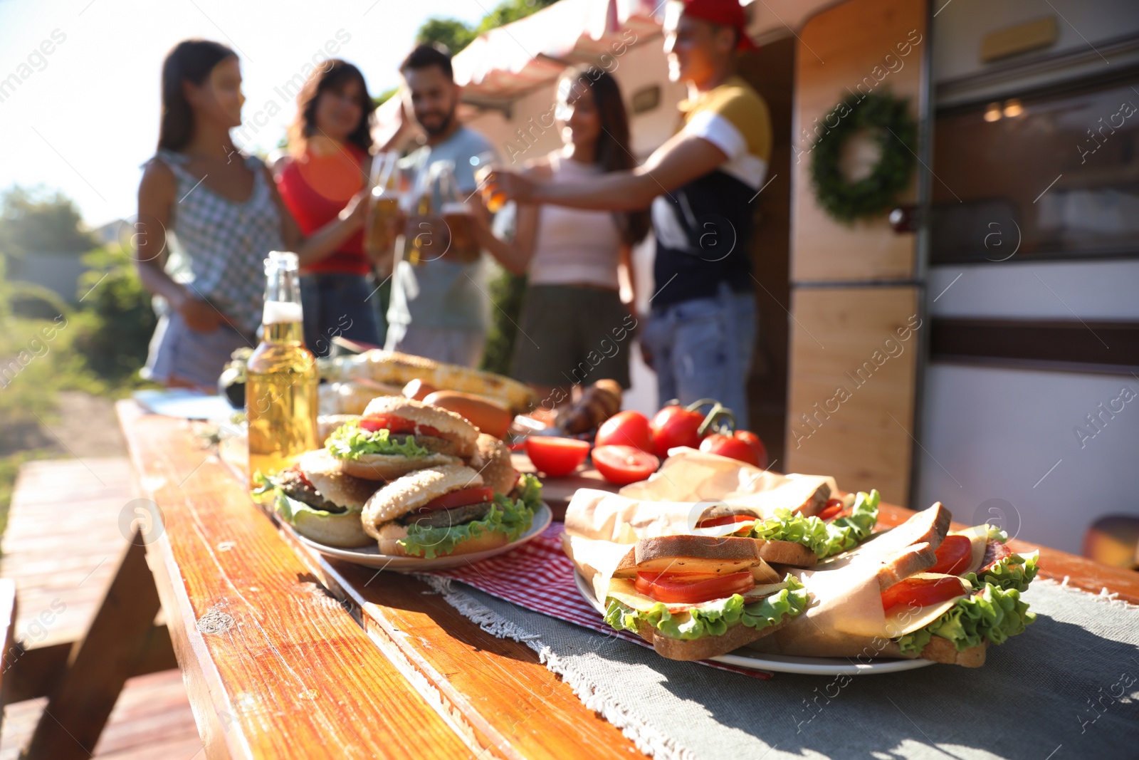 Photo of Friends toasting with bottles of drinks near motorhome, focus on table full of tasty food. Camping season