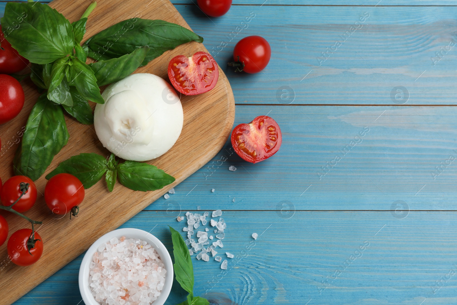 Photo of Delicious burrata cheese with basil and tomatoes on light blue wooden table, flat lay. Space for text