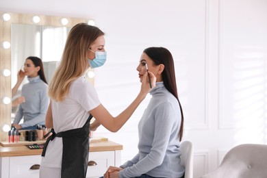 Photo of Makeup artist in protective mask working with woman indoors. Preventive measures during COVID-19 pandemic