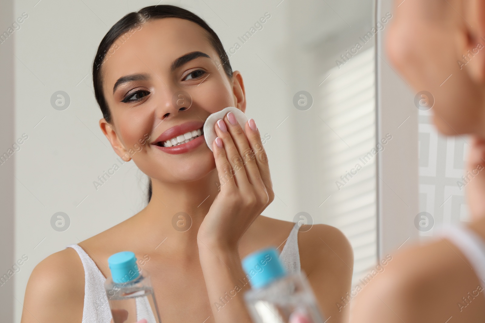 Photo of Beautiful woman removing makeup with cotton pad near mirror indoors