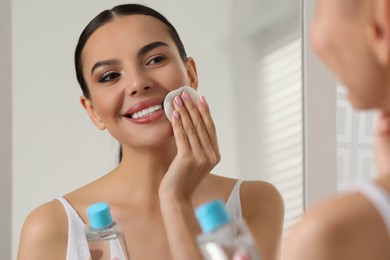 Beautiful woman removing makeup with cotton pad near mirror indoors