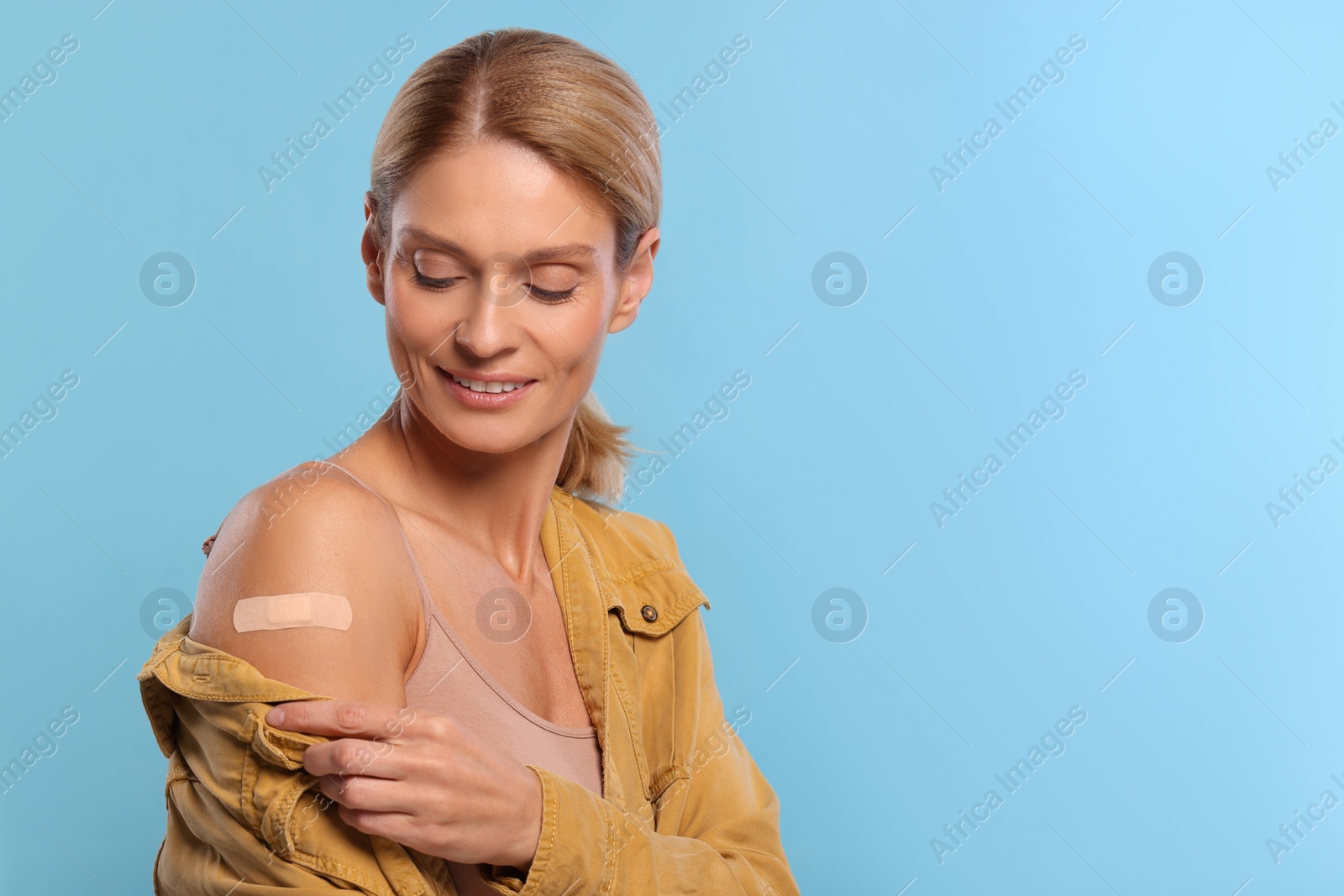 Photo of Smiling woman with adhesive bandage on arm after vaccination on light blue background. Space for text